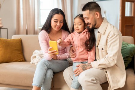 Foto de Primer Gadget. Los padres asiáticos y la hija del bebé usando el teléfono inteligente se divierten juntos en línea sentados en el sofá en casa. Familia de tres navegando por la web disfrutando de la tecnología y las innovaciones - Imagen libre de derechos