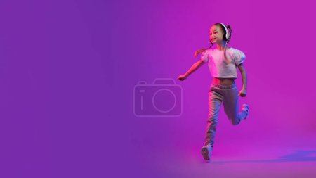 Téléchargez les photos : Joyeux écolier mignon petite fille utilisant des écouteurs stéréo sans fil modernes dans la lumière lumineuse au néon, courant vers l'espace de copie. Energie vibrante et rythme de bonne musique de qualité, bannière web - en image libre de droit