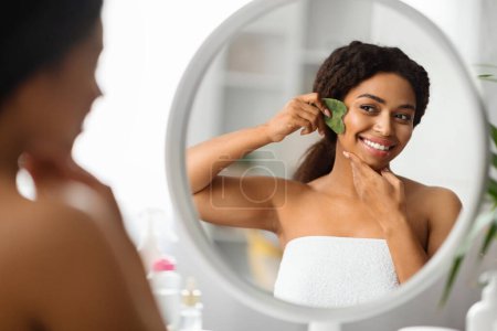 Téléchargez les photos : Belle jeune femme noire faisant massage facial avec gua sha outil, souriant attrayant visage féminin afro-américain massant tout en se tenant près du miroir à la maison, faire des soins de beauté après le bain - en image libre de droit