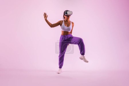 Téléchargez les photos : Améliorer la condition physique grâce à la réalité vitale. Femme noire active avec lunettes VR dansant et explorant la technologie 3D sur fond de lumière rose fluo, pleine longueur - en image libre de droit