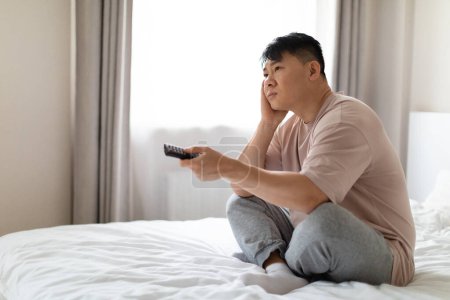 Téléchargez les photos : Malheureux ennuyé homme asiatique d'âge moyen portant un pyjama assis sur le lit à la maison, tenant la télécommande et touchant son visage, regardant la télévision, à la recherche d'un beau film ou d'une émission de télévision, copier l'espace - en image libre de droit