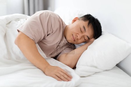 Téléchargez les photos : Seul bel homme asiatique d'âge moyen portant un pyjama dormant paisiblement seul dans son lit à la maison, profitant de son lit confortable, matelas orthopédique et oreiller. Concept de sommeil sain - en image libre de droit
