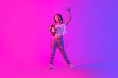Foto de Retrato de alegre niña juguetona y elegante cantando sobre fondo futurista rosa. Joven cantante con micrófono y auriculares inalámbricos. Infancia, concepto de pasatiempos para niños - Imagen libre de derechos
