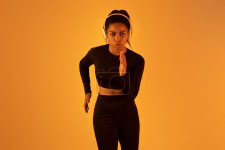 Photo for Sporty young african american woman running or jogging towards camera, ready for sprint, wearing headset on orange neon studio background. Cardio workout and competition concept. - Royalty Free Image