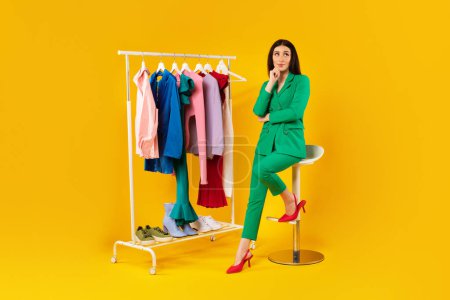 Photo for Pretty stylist lady sitting near garment rack and choosing new clothes, woman thinking about creating new outfits on yellow studio background, full length, copy space - Royalty Free Image