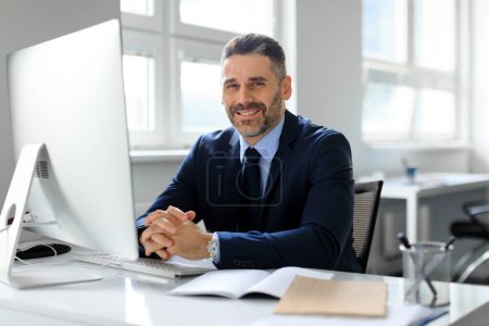 Téléchargez les photos : Portrait d'un homme d'affaires d'âge moyen heureux assis devant l'ordinateur sur le lieu de travail au bureau, souriant à la caméra. Entrepreneur assis au bureau. Entreprises et entrepreneuriat - en image libre de droit
