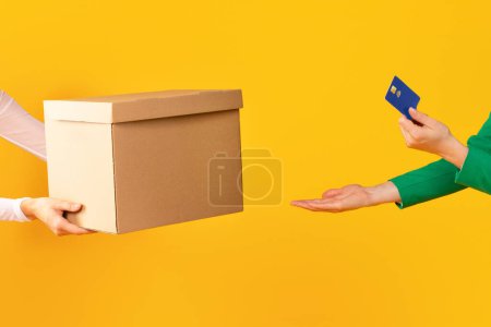 Téléchargez les photos : Les mains féminines tenant la carte de crédit et prenant la boîte en carton du transporteur ou du vendeur sur fond jaune studio, livraison, emballage ou concept d'achat de paiement - en image libre de droit