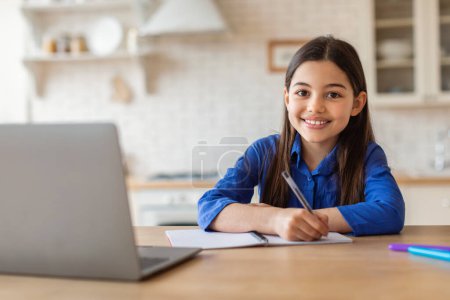 Foto de Educación en línea. Chica joven y feliz tomando notas en el ordenador portátil abrazando el aprendizaje electrónico, estudiando desde casa, sentado en el escritorio haciendo la tarea de la escuela. Tiro de colegiala exitosa durante la clase digital - Imagen libre de derechos