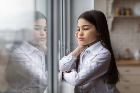 Photo for Kid Depression. Unhappy Preteen Girl Looking Out Of Window Suffering From Unhappiness And Loneliness Standing At Home. Schoolgirl Posing Alone Indoors. Child Mental Health Issues Concept - Royalty Free Image