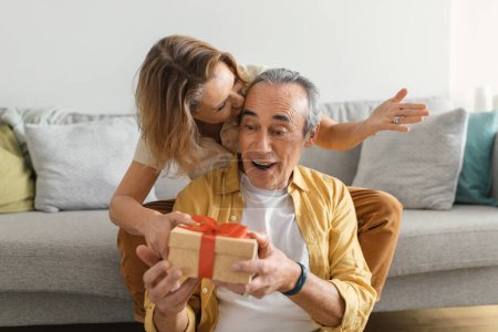 Téléchargez les photos : Bonne femme âgée donnant boîte cadeau à son mari, félicitant avec anniversaire, assis sur le canapé à la maison. Fête, anniversaire, émotions des gens et surprise - en image libre de droit