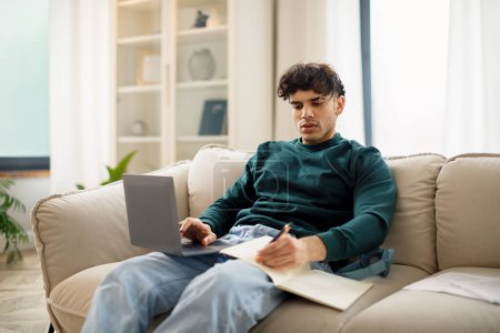 Téléchargez les photos : E-Learning. Étudiant arabique concentré gars travaillant avec ordinateur portable et prendre des notes assis sur le canapé dans le salon moderne. Homme naviguant sur Internet et écrivant se préparant aux examens à la maison - en image libre de droit