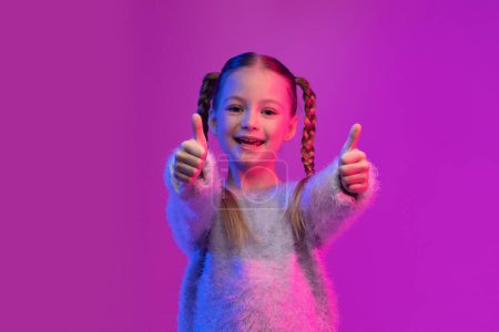 Photo for I like it. Happy cutie preteen girl with braids in stylish sweater showing thumb ups and smiling on colorful background in luminous neon light, kid recommend something cool, nice - Royalty Free Image