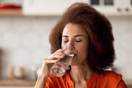 Foto de Joven mujer afroamericana bebiendo agua mineral limpia de vidrio en casa, mujer negra sedienta disfrutando de una bebida saludable, cuidando la hidratación corporal, primer plano con espacio libre - Imagen libre de derechos