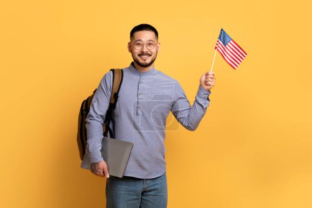 Téléchargez les photos : Concept d'échange d'étudiants. Jeune homme asiatique tenant l'ordinateur portable et le drapeau américain des États-Unis, heureux homme millénaire regardant et souriant à la caméra tout en se tenant sur fond jaune studio, Copier l'espace - en image libre de droit