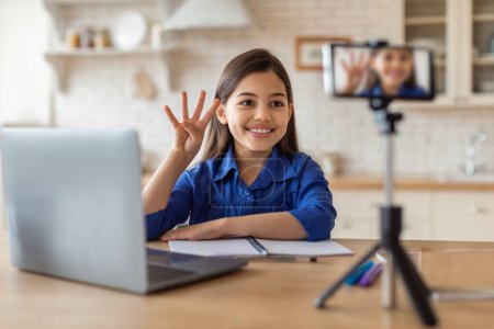 Photo for Digital Schooling. Happy School Girl Having Online Class Via Video Call On Phone, Showing Four To Smartphone On Tripod Sitting At Laptop At Home. Successful E-Learning. Selective Focus - Royalty Free Image