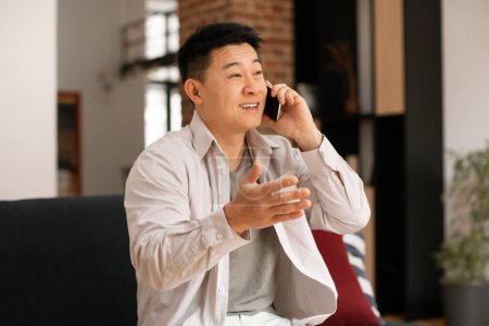 Photo for Emotional middle aged asian man in casual outwear sitting on sofa, having phone conversation with friend or lover at home, looking aside and gesturing - Royalty Free Image