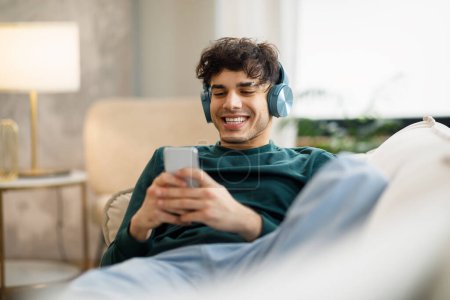 Photo for Cheerful Middle Eastern Guy Websurfing On Smartphone Wearing Wireless Headphones, Having Fun Relaxing On Sofa At Home. Young Man Listening To Music Online Via Musical App. Selective Focus - Royalty Free Image