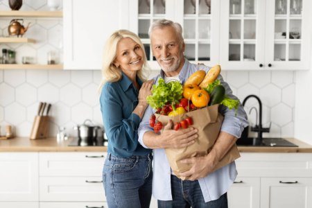 Foto de Feliz pareja de ancianos posando con la bolsa de comestibles en el interior de la cocina, los cónyuges jubilados ancianos sosteniendo paquete con verduras frescas en casa, mirando y sonriendo a la cámara, disfrutando de la vida en la jubilación - Imagen libre de derechos