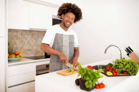 Téléchargez les photos : Cuisine Maestro. Joyeux cuisinier afro-américain portant tablier coupe légumes pour salade fraîche profiter de la cuisine Loisirs à la maison, souriant à la caméra. Concept de recettes saines - en image libre de droit