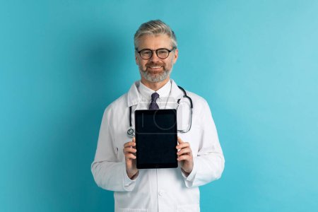 Photo for Telemedicine and Virtual Consultations. Handsome middle aged doctor showing digital tablet with mockup to conduct remote appointments, diagnose patients, provide medical advice, blue background - Royalty Free Image