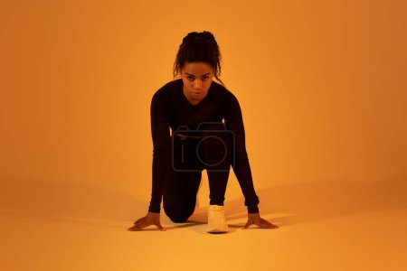 Photo for Young african american runner woman doing crouch start running on orange neon studio background, wearing black fitwear and looking at camera - Royalty Free Image
