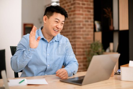Téléchargez les photos : Souriant asiatique d'âge moyen homme d'affaires agitant la main à la webcam ordinateur portable, ayant réunion en ligne à la maison. Salut, salut et bonjour, appel vidéo, travail d'étude et affaires à distance - en image libre de droit