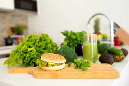 Téléchargez les photos : Detox contre la malbouffe. Burger et verre de Smoothie vert debout sur la table à manger parmi les légumes et les herbes dans la cuisine moderne intérieure. Concept de choix d'aliments sains ou malsains - en image libre de droit
