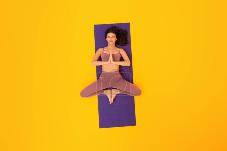 Foto de Mindful Fitness. Vista superior de la mujer en forma que medita haciendo yoga con los ojos cerrados que mienten en la estera del ejercicio que usa Fitwear en fondo amarillo del estudio. Concepto de relajación y tranquilidad - Imagen libre de derechos
