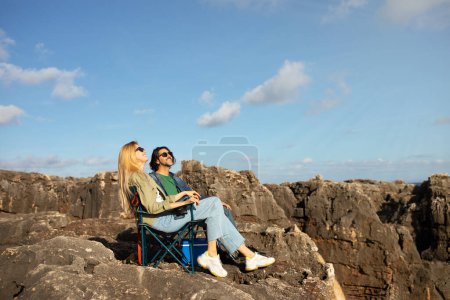 Photo for Happy Young Couple Relaxing In Camping Chairs On Rocky Coast, Cheerful Romantic Man And Woman Sunbathing And Smiling, Enjoying Outdoor Picnic On Rocks, Having Date Outside On Sunny Day, Copy Space - Royalty Free Image