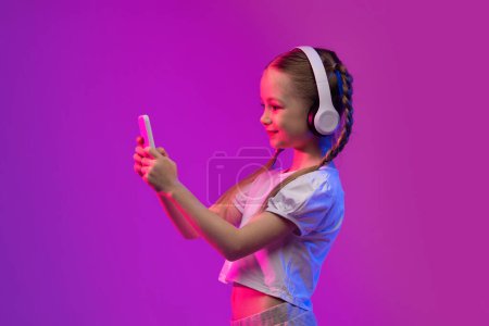 Téléchargez les photos : Joyeux adorable préadolescente avoir un appel vidéo avec des amis, en utilisant des écouteurs sans fil et téléphone portable sur fond coloré dans la lumière au néon, souriant à l'écran du smartphone, copier l'espace - en image libre de droit