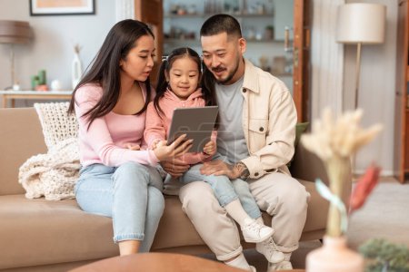 Foto de Familia y Gadgets. Padres chinos e hijita usando Tablet Computer Websurf juntos en casa. Papi, mami y el niño que participan en la diversión y la actividad de Internet juntos en fin de semana - Imagen libre de derechos