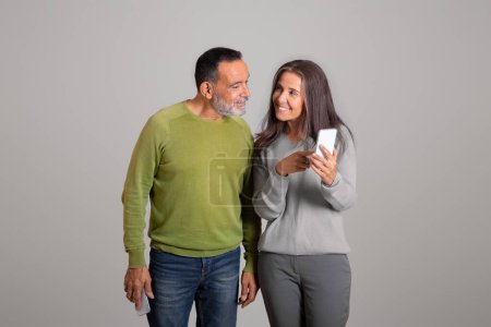 Photo for Glad caucasian aged woman shows phone to man, chatting in social network, recommends ad and offer, isolated on gray background, studio. Blog, communication remotely, app banking - Royalty Free Image