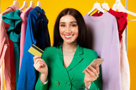 Foto de Mujer caucásica emocionada usando el teléfono celular y la tarjeta de crédito para ir de compras, posando cerca del estante de la prenda y sonriendo a la cámara sobre el fondo amarillo del estudio - Imagen libre de derechos