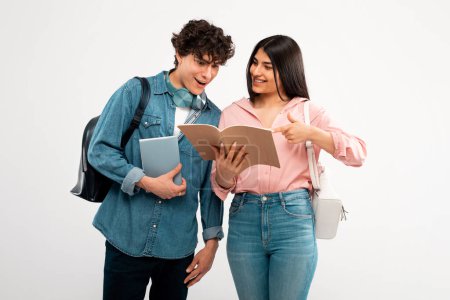 Photo for College Friends. Cheerful Student Lady Showing Her Workbook To Excited Guy With Backpack, Learning Together Over White Background. Studio Shot Of Enthusiastic Fellow Students Discussing Homework - Royalty Free Image