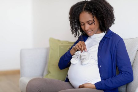 Photo for Glad curly young black woman in domestic clothes with big belly enjoy pregnancy, hold small shoes in living room interior. Health care, gender party, motherhood and expectation of child at home - Royalty Free Image