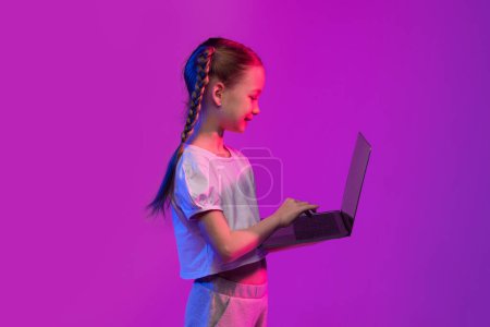 Photo for Virtual learning, online education, e-schooling concept. School aged child cute little girl with braids typing on modern pc laptop keyboard over colorful luminous background, copy space, side view - Royalty Free Image