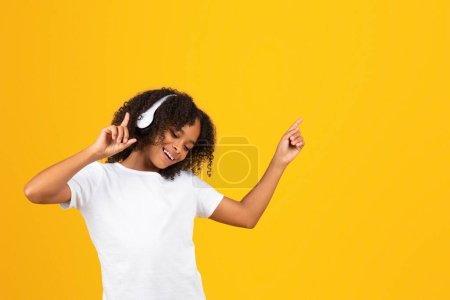 Photo for Glad curly teen african american girl in white t-shirt and wireless headphones dancing, listen music, enjoy free time isolated on yellow studio background. Audio app for relax, fun - Royalty Free Image