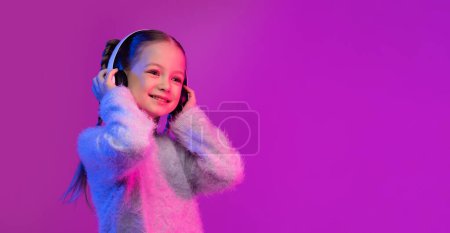 Téléchargez les photos : Joyeux enfant jolie fille de préadolescence aux cheveux blonds en utilisant de nouveaux écouteurs sans fil blancs, en regardant l'espace de copie et en souriant sur fond de studio rose dans la lumière au néon, en écoutant de la musique, bannière web - en image libre de droit
