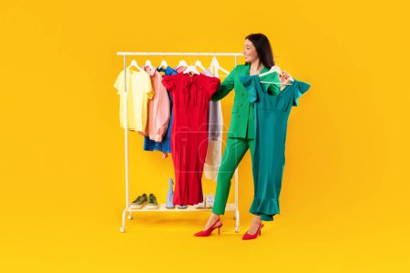 Photo for Fashion and trends concept. Excited shopaholic lady choosing between two elegant dresses, standing near clothing rail and buying new outfit over yellow studio background, full length - Royalty Free Image