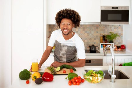 Photo for Culinary Master. Shot Of Happy Black Man Posing Near Kitchen Countertop With Fresh Food Products And Vegetables, Cooking Salad For Dinner, Enjoying Tasty Healthy Food Preparation - Royalty Free Image