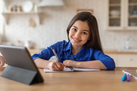 Photo for E-Learning. Happy Schoolgirl Attending School Through Digital Tablet From Home, Learning And Doing Homework Online Taking Notes Near Computer, Smiling To Camera Posing Indoors - Royalty Free Image