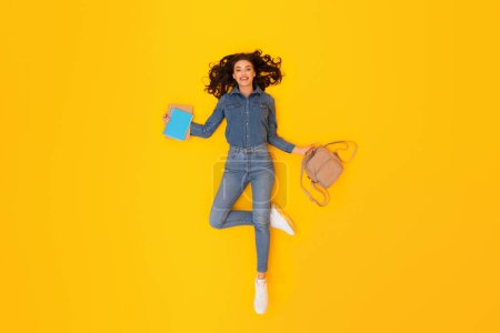 Photo for Ready for Knowledge. Glad Woman Student Holding Backpack And Study Books Expressing Positive Emotions And Learning Anticipation Lying On Yellow Studio Backdrop. Above View - Royalty Free Image