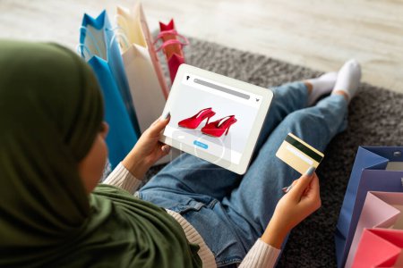 Foto de Mujer árabe joven en hijab usando tarjeta de crédito y tableta con tienda de moda en la pantalla, ofreciendo sitio web o aplicación de compras, sentado en el suelo en casa con bolsas de comprador. Entrega de zapatos y finanzas - Imagen libre de derechos