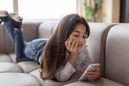 Photo for Digital Depression. Unhappy Preteen Girl Using Smartphone Reading Depressing News And Suffering From Lack Of Communication Lying On Sofa At Home. Gadgets Technology And Negative Emotions - Royalty Free Image