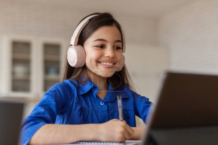 Foto de Oferta de E-Learning. Chica del niño de la escuela feliz usando el ordenador portátil que usa los auriculares, viendo la conferencia virtual en casa. Colegiala participando en el aprendizaje en línea en el escritorio interior. Conocimiento digital y estudio remoto - Imagen libre de derechos