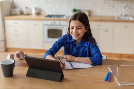 Foto de Aprendizaje electrónico. Happy Middle Eastern Schoolgirl Using Digital Tablet And Taking Notes Embracing Online Education At Home (en inglés). Niña haciendo la tarea en línea Excelling en sus estudios remotos - Imagen libre de derechos