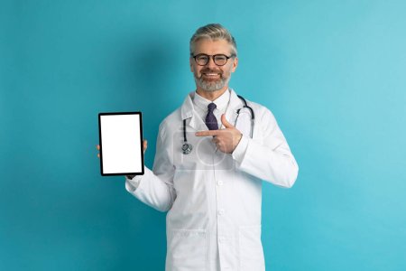 Photo for Technology and Communication in Healthcare. Cheerful mature doctor showing digital tablet with white empty screen on blue background, communicate with colleagues, patients, or medical staff, mockup - Royalty Free Image