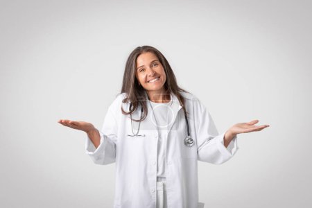 Foto de Mujer doctora con estetoscopio que tiene dudas, extendiendo las manos a un lado y levantando hombros, mirando y sonriendo a la cámara sobre fondo gris. Concepto incierto - Imagen libre de derechos