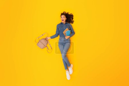 Photo for Academic Joy. Top View Of Enthusiastic Student Woman Grasping Backpack And Study Materials Smiling Towards Camera Lying On Yellow Studio Backdrop, Full Length Shot. Modern University Study Concept - Royalty Free Image