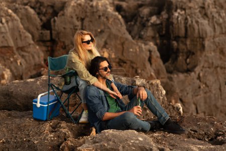 Téléchargez les photos : Portrait de romantique jeune couple ayant pique-nique en plein air sur les rochers et embrasser, heureux amants millénaire se détendre dans les chaises de camping et boire de la bière, homme et femme regardant coucher de soleil, Copier l'espace - en image libre de droit